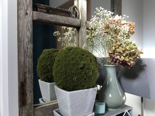 Bouquet d’hortensias et de petites fleurs blanches dans un vase vert grisé, près d’une boule de mousse devant un miroir en bois patiné.